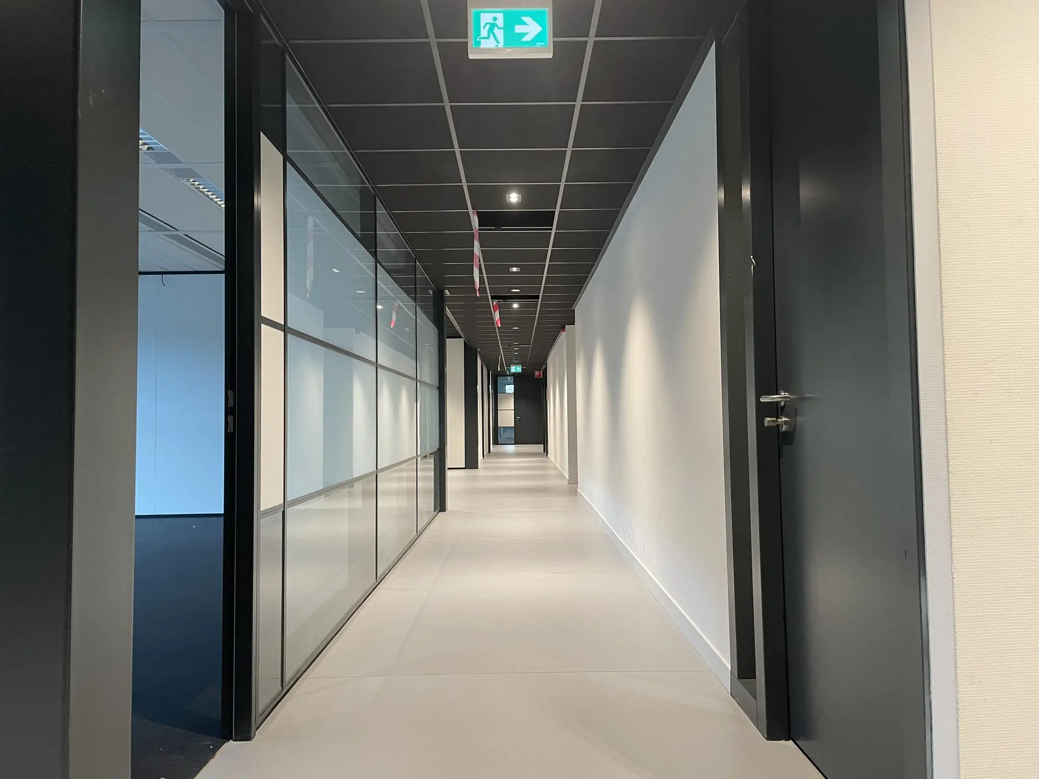 Lange, moderne kantoorhal met zwarte deuren, glazen wanden en een nooduitgang bovenaan.