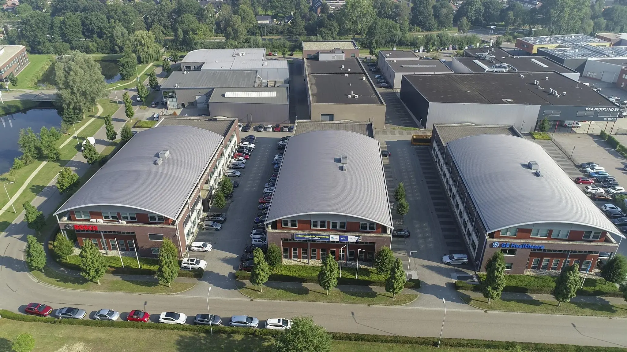 Luchtfoto van drie moderne bedrijfsgebouwen aan de De Wel in een groen bedrijvenpark, met zicht op parkeergelegenheid en omliggende kantoren.