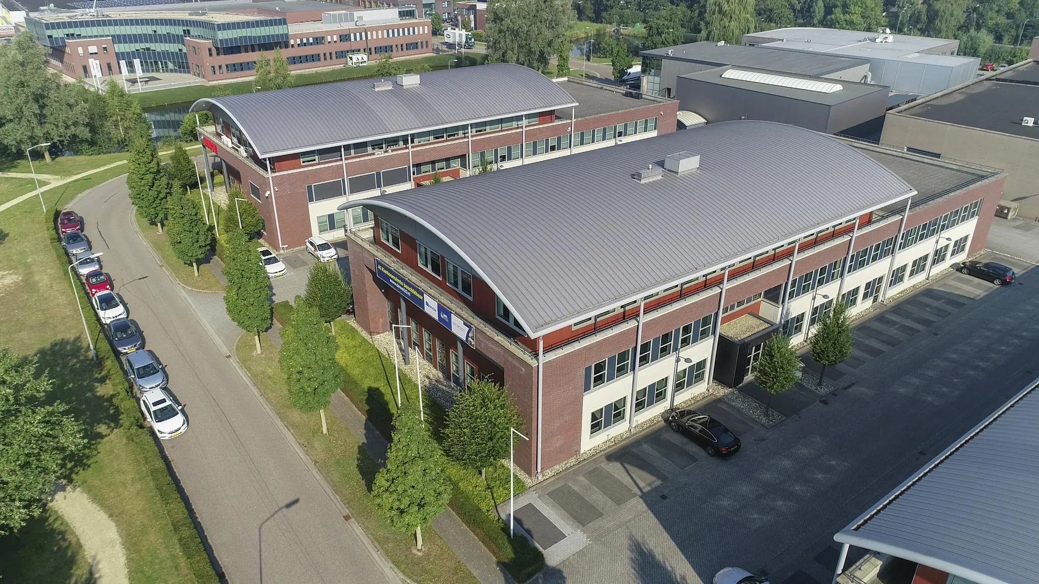 Luchtfoto van het bedrijfsverzamelgebouw De Wel met omliggende kantoren en geparkeerde auto's in een groen bedrijfspark.