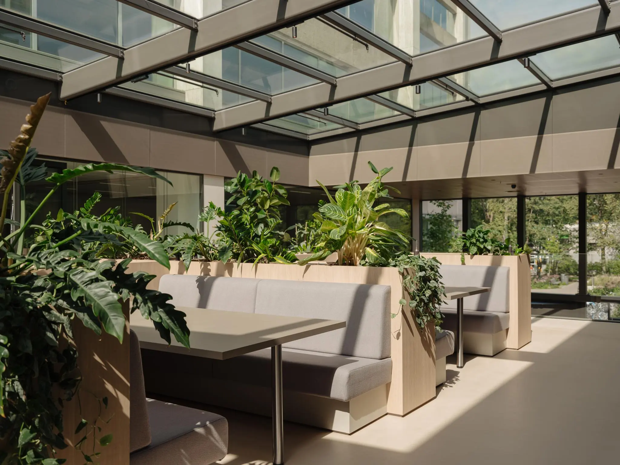 Moderne zithoek met grijze banken en houten plantenbakken onder een glazen dak in een licht atrium aan de Nachtwachtlaan.