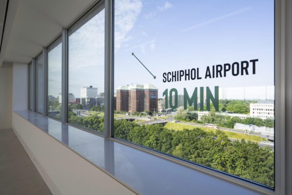 Uitzicht vanuit een kantoorgebouw aan de Nachtwachtlaan op de Amsterdamse Zuidas met tekst op het raam die aangeeft dat Schiphol Airport in 10 minuten bereikbaar is.