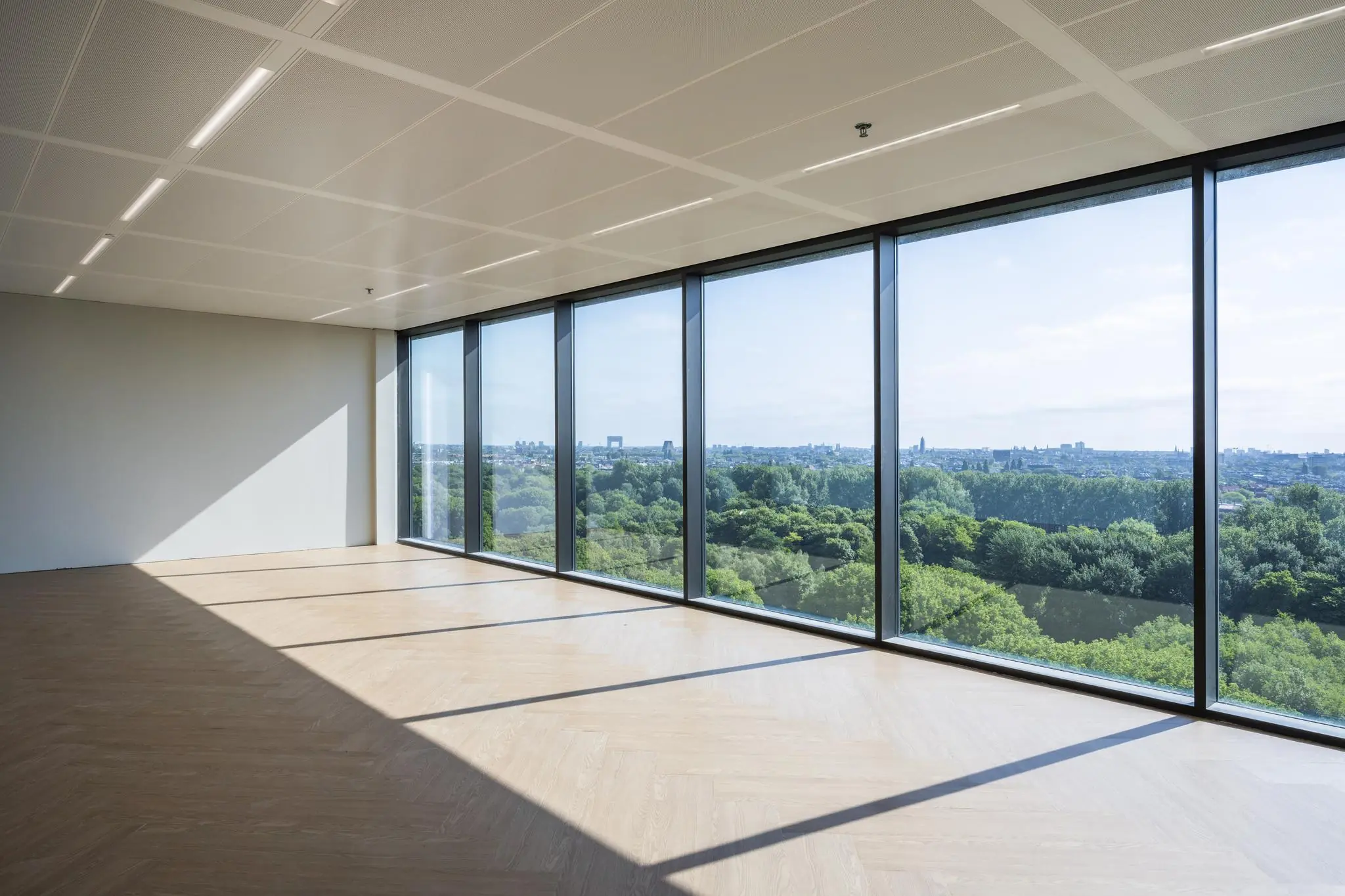 Lichte, lege kantoorruimte met houten vloer en grote ramen met uitzicht op het groen en de skyline vanaf de Nachtwachtlaan.