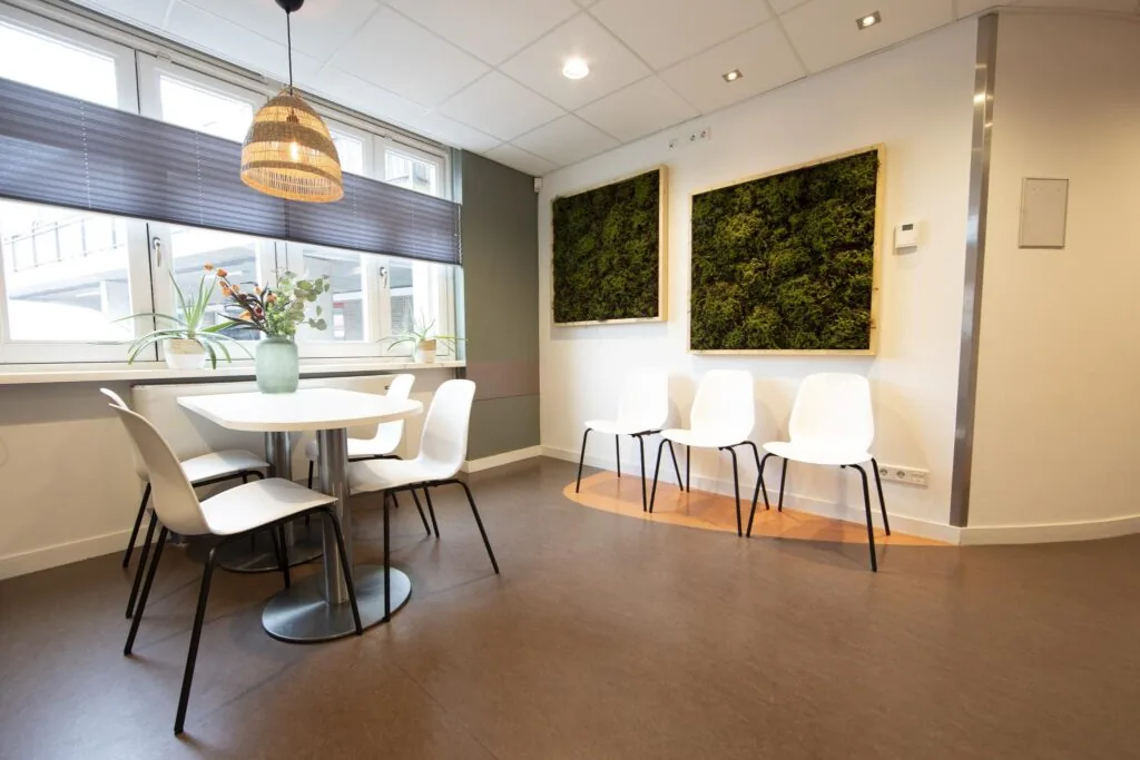 Wachtkamer met witte stoelen, een ronde tafel, planten en twee groene moskunstwerken aan de muur.