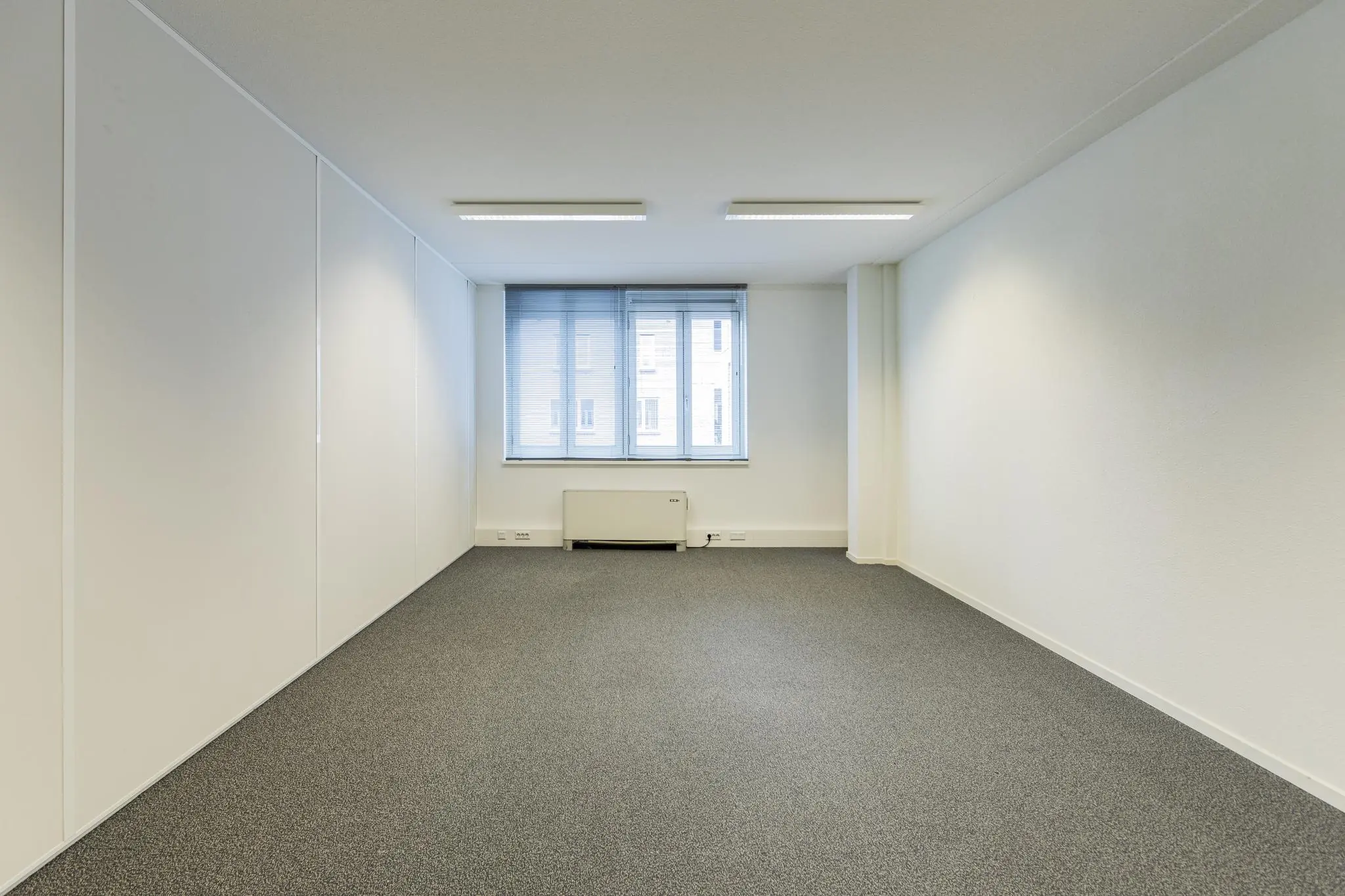 Leege, rechthoekige kantoorruimte in het gebouw Middenburcht met witte muren, grijs tapijt en een raam met jaloezieën.