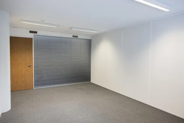 Lege kantoorruimte in De Middenburcht met witte wanden, grijs tapijt en gesloten jaloezieën voor een glazen wand.
