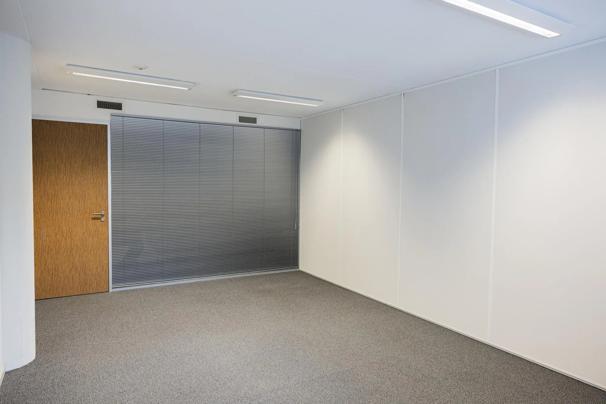Lege kantoorruimte in De Middenburcht met witte wanden, grijs tapijt en gesloten jaloezieën voor een glazen wand.