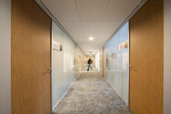 Verlichtte kantoorhal van kantoorgebouw Middenburcht met houten deuren en glazen wanden.