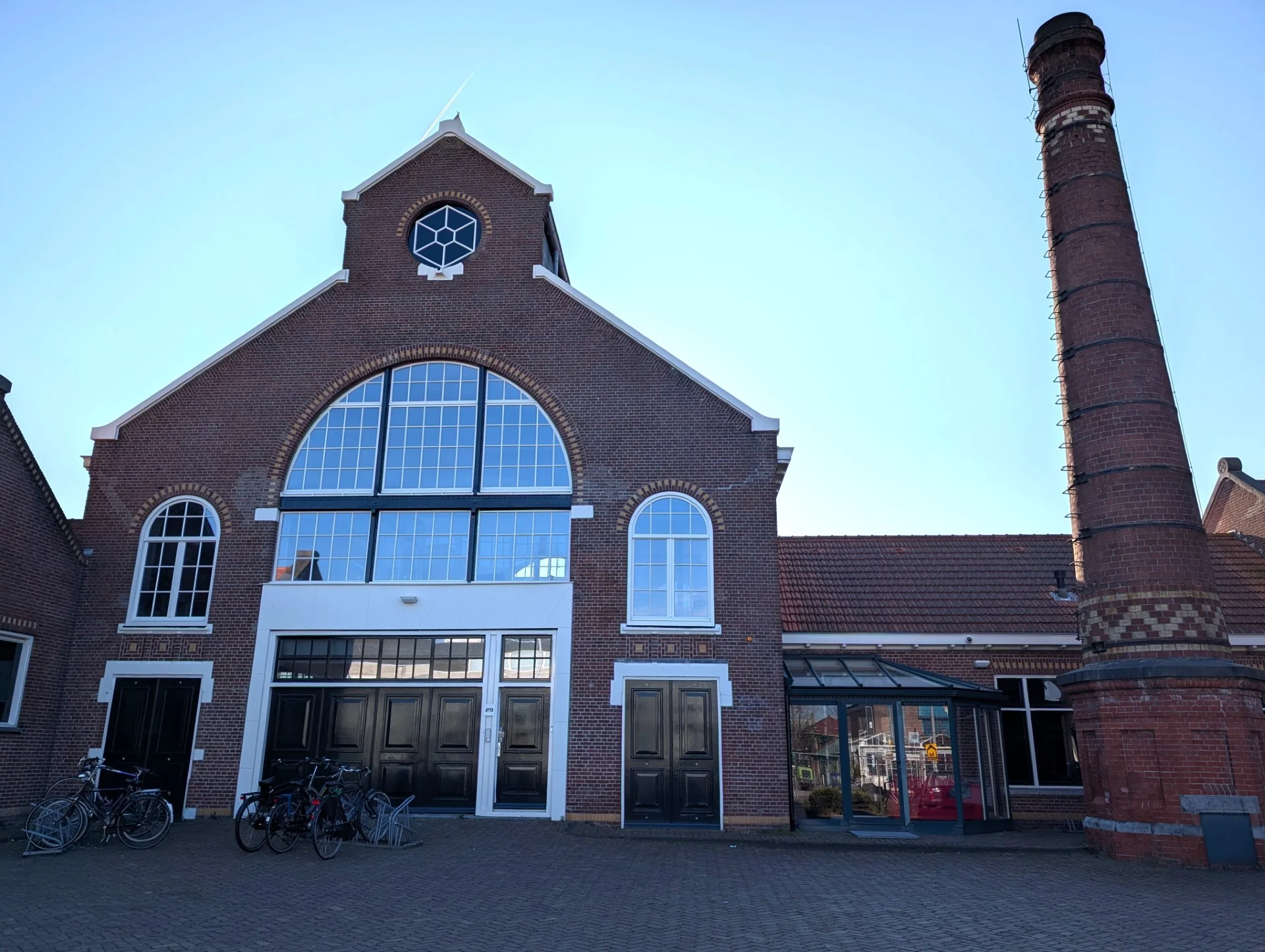 Vooraanzicht van de monumentale bakstenen fabriek aan het Gedempt Hamerkanaal met grote ramen, fietsenrekken en een opvallende schoorsteen.