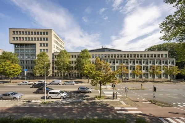 Straatbeeld van de Derkinderenstraat met een groot kantoorgebouw, bomen en passerende auto's op een zonnige dag.