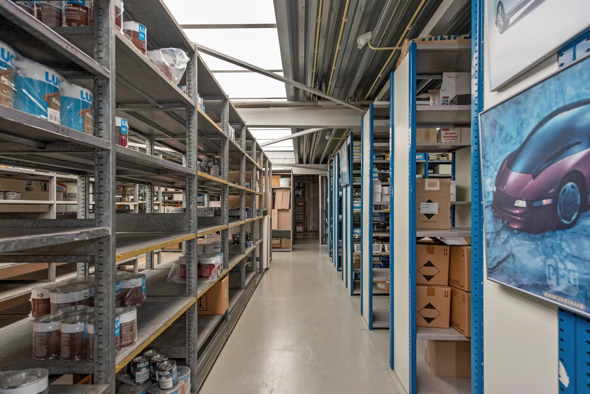 Gangpad in een magazijn aan de Lagosweg met metalen stellingen gevuld met verfblikken, dozen en auto-onderdelen.