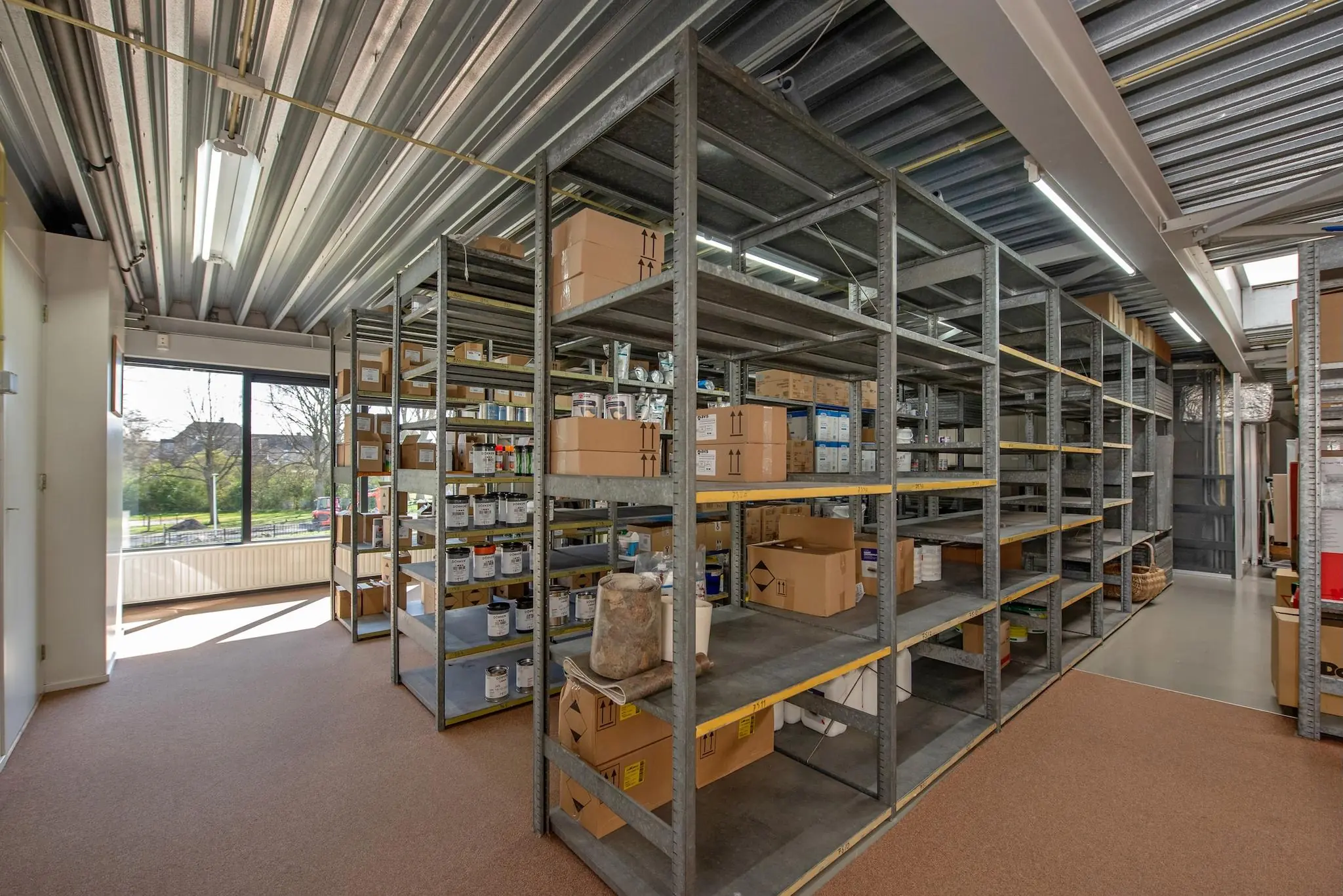 Opslagruimte aan de Lagosweg met metalen stellingen vol dozen, verfblikken en kantoorartikelen.
