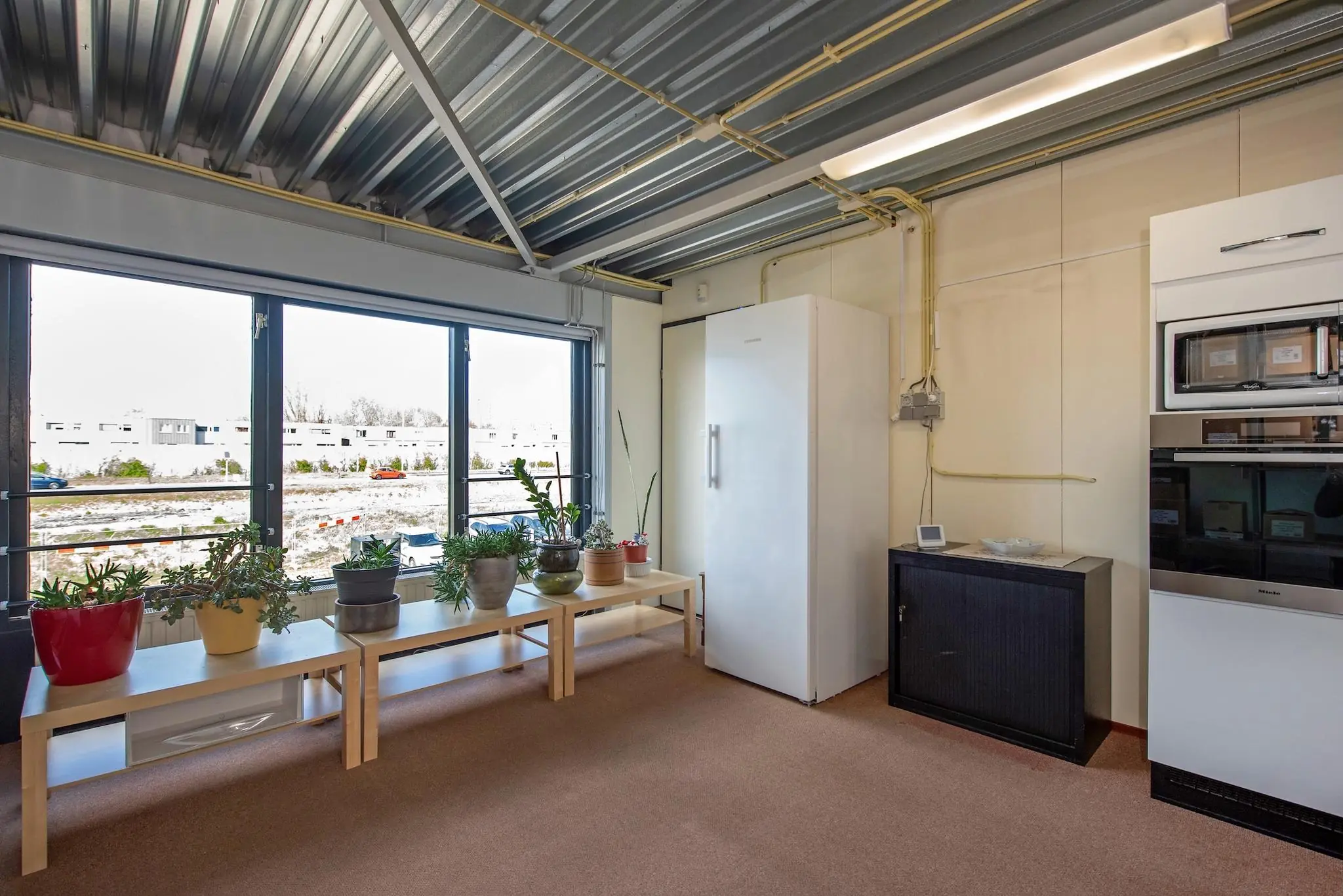 Kantoorkeuken aan de Lagosweg met grote ramen, kamerplanten, een koelkast en ingebouwde keukenapparatuur.