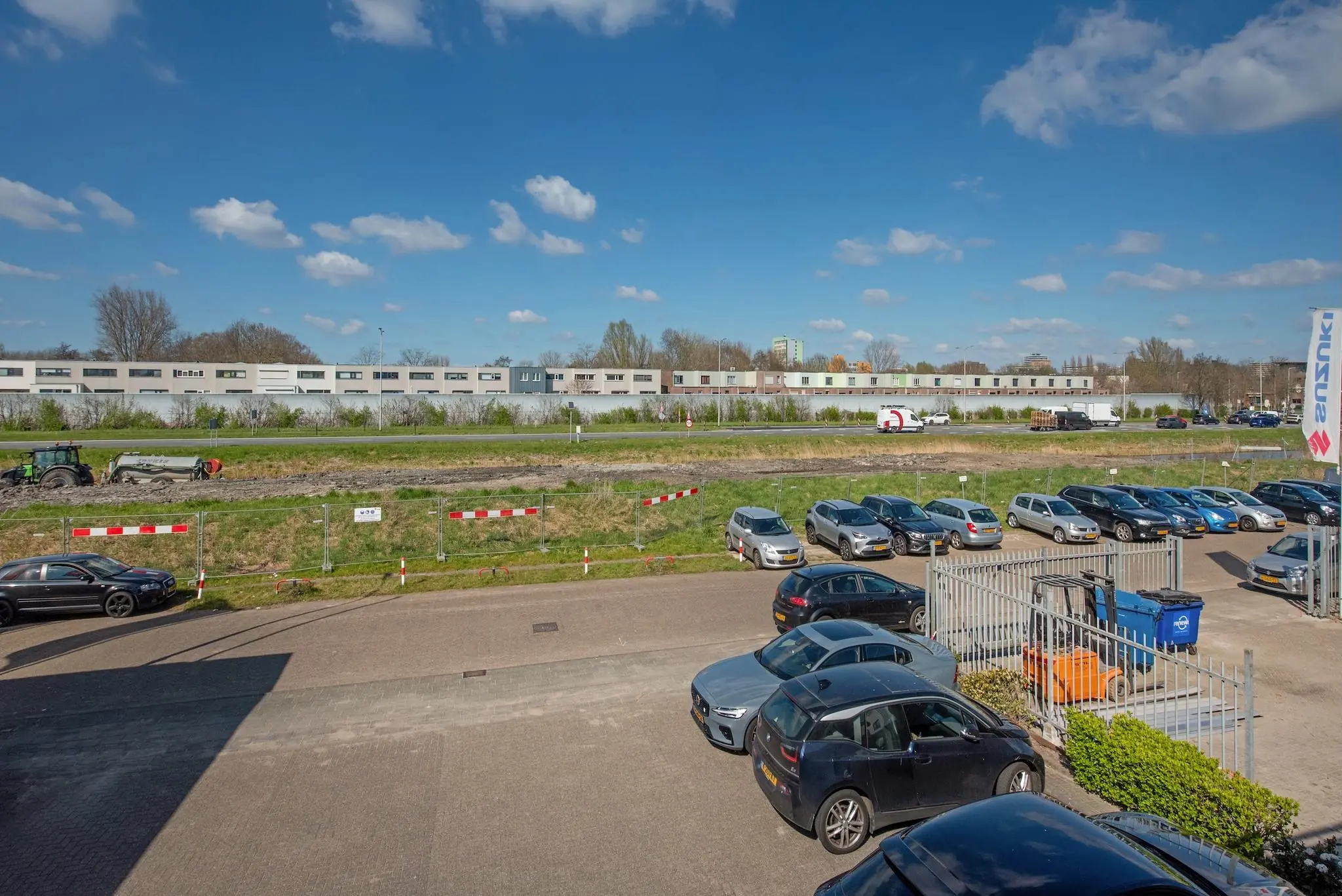 Parkeerterrein aan de Lagosweg met geparkeerde auto's, een tractor op een modderig terrein en een lang wit gebouw op de achtergrond.