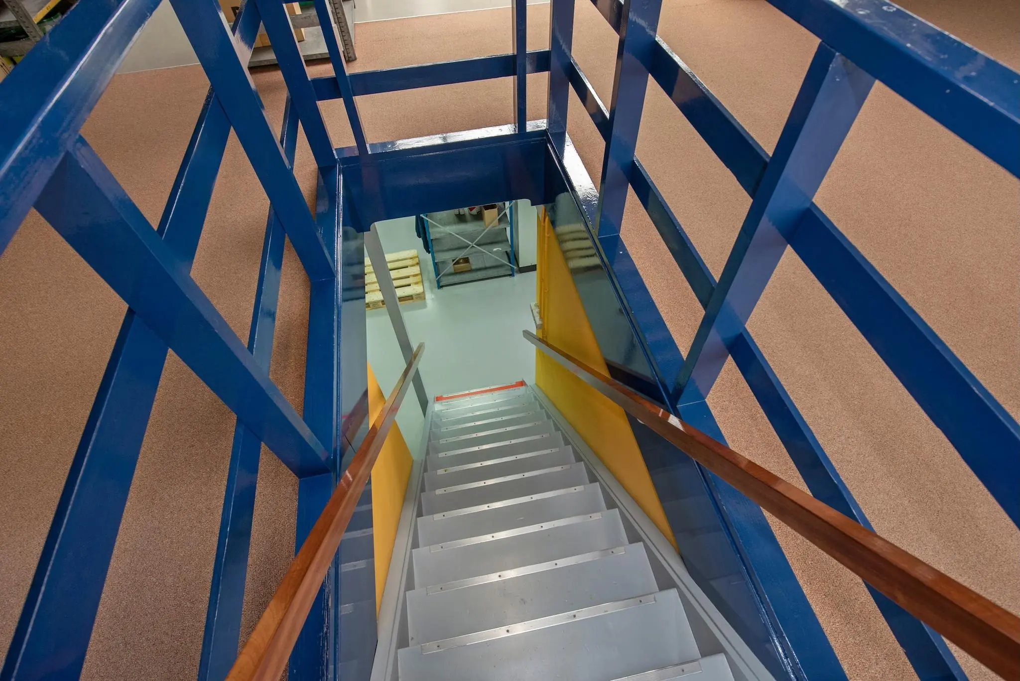 Blik van bovenaf op een metalen trap met blauwe leuningen en gele zijwanden in een industrieel gebouw aan de Lagosweg.
