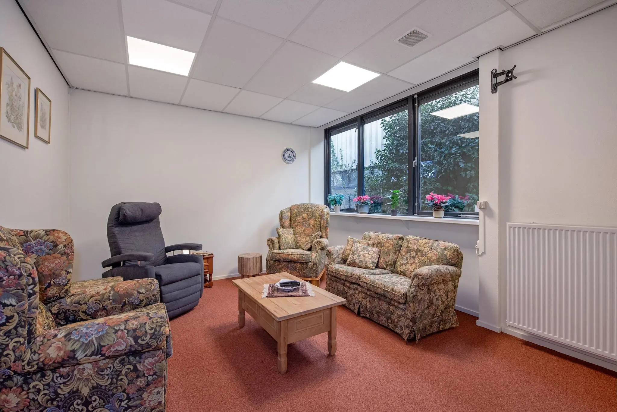 Kleine zithoek met bloemenpatronen meubilair, houten salontafel en grote ramen met planten op de vensterbank.