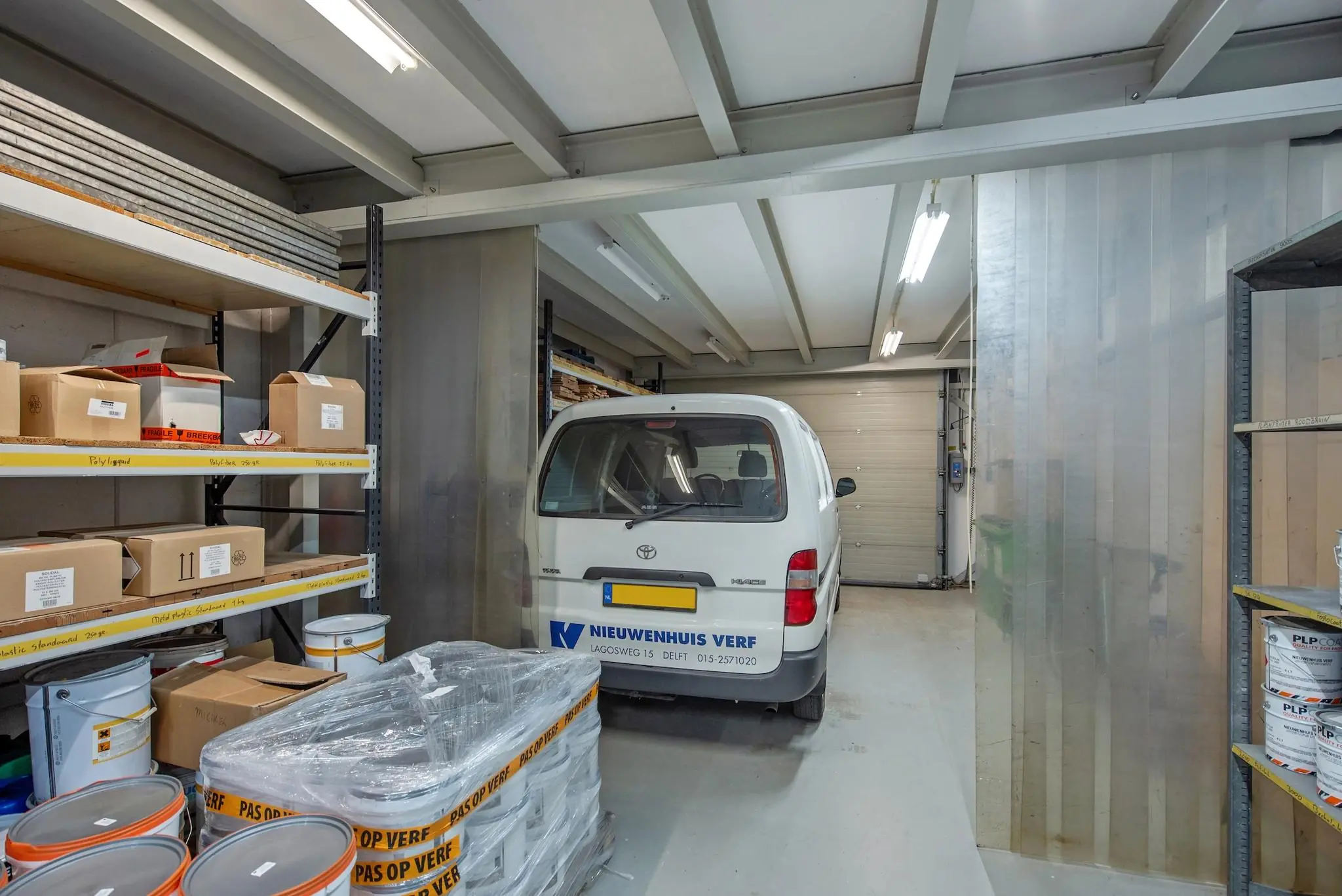 Magazijnruimte met een bedrijfsauto van Nieuwenhuis Verf, omringd door verfblikken en dozen op stellingen.