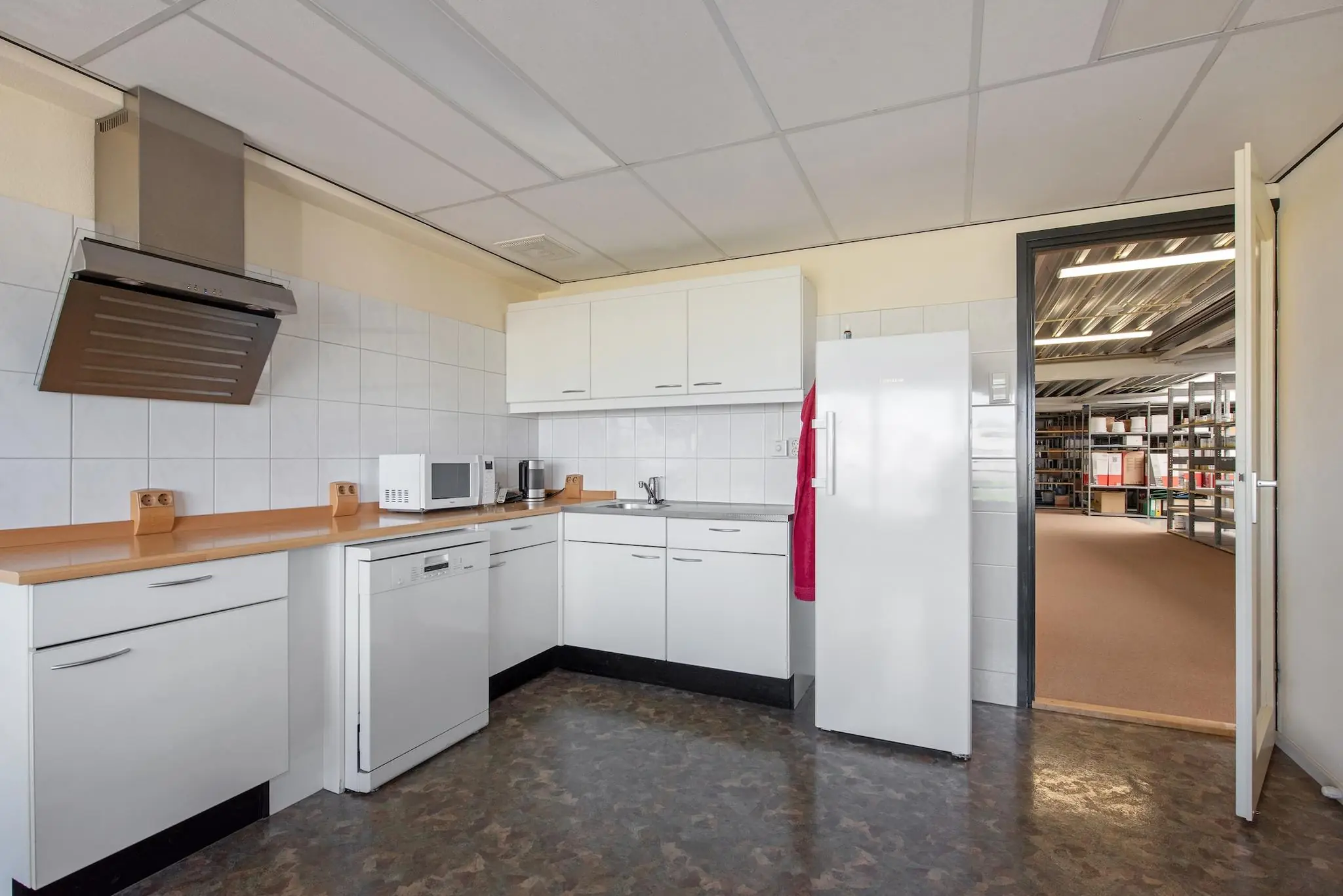 Kleine bedrijfskeuken met witte kasten, apparatuur en zicht op een aangrenzende opslagruimte met archiefrekken.