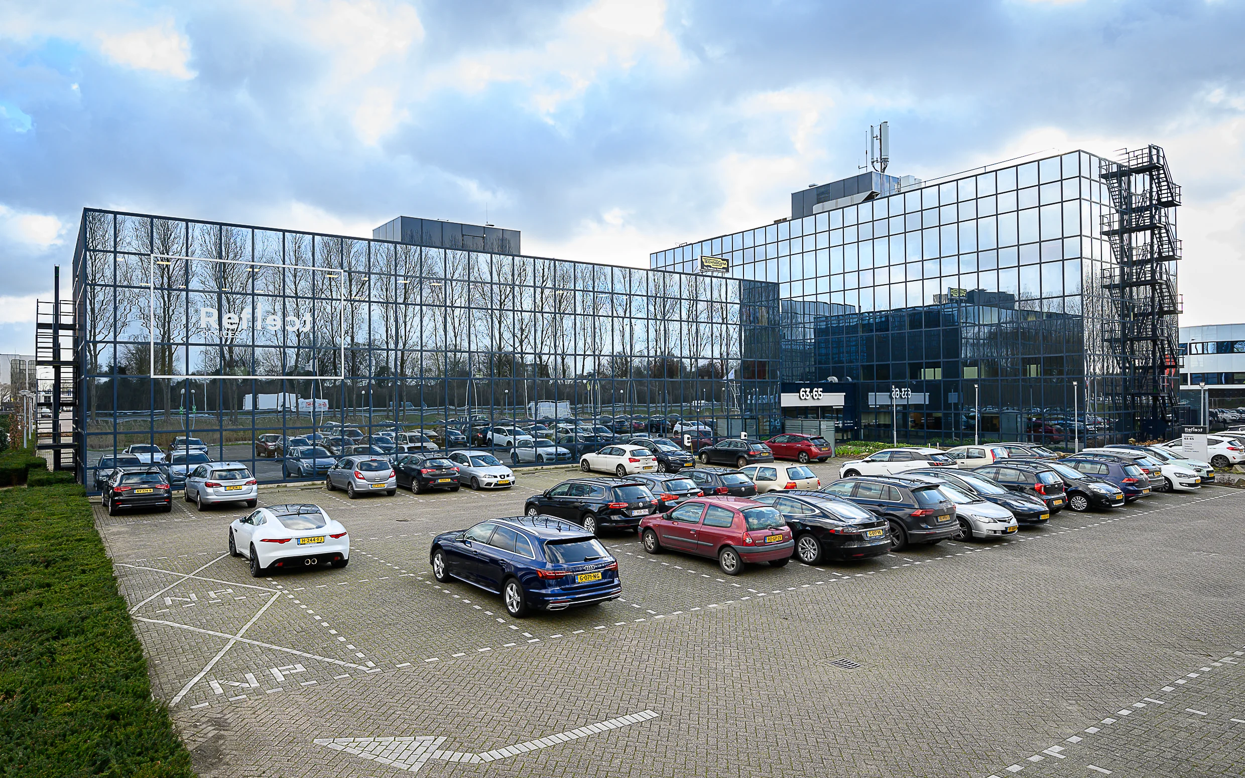 Modern glass office building at Essebaan 63-65 with a full parking lot in front.