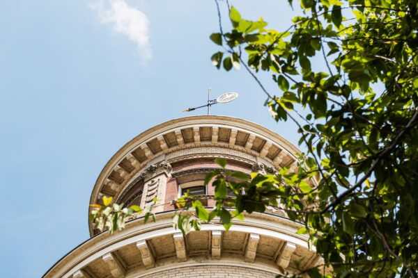 Rond torentje van een historisch gebouw aan de Leidseweg, deels omringd door groene bladeren tegen een blauwe lucht.
