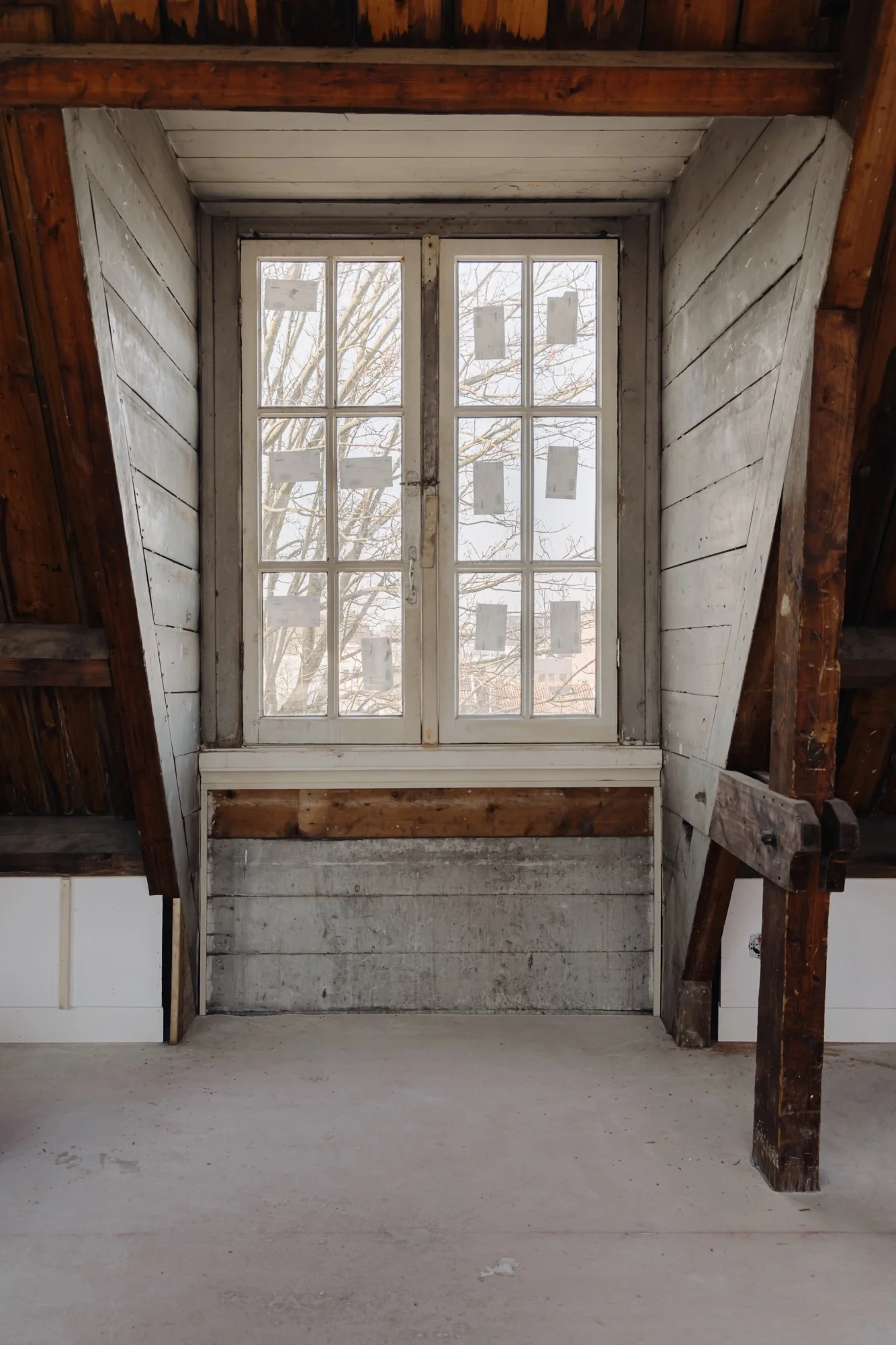 Houten zolderruimte met een oud raam aan de Leidseweg en zicht op een boom en gebouw buiten.