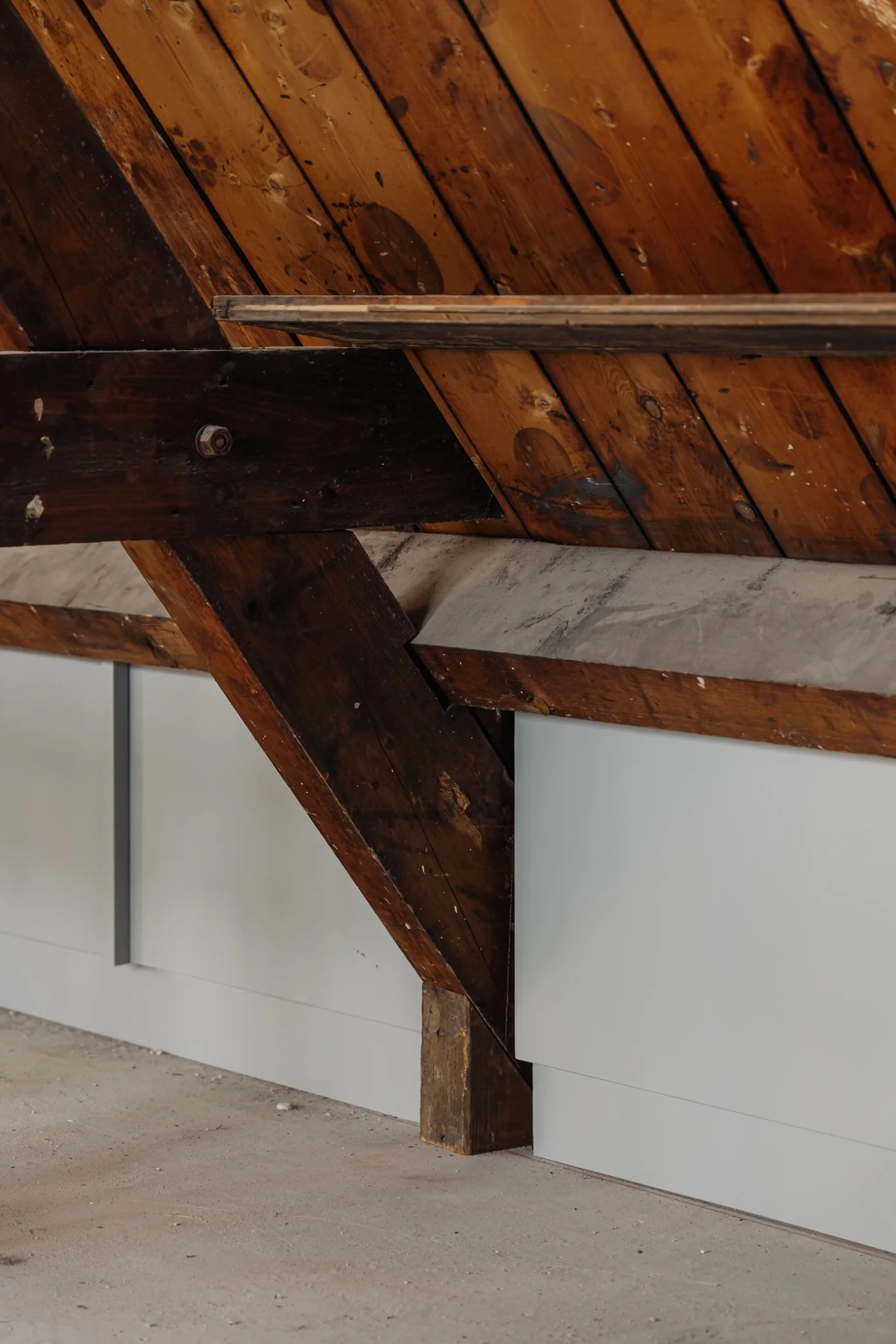Houten draagbalkconstructie in een schuine zoldermuur aan de Leidseweg met wit afgewerkte wandpanelen en betonnen vloer.