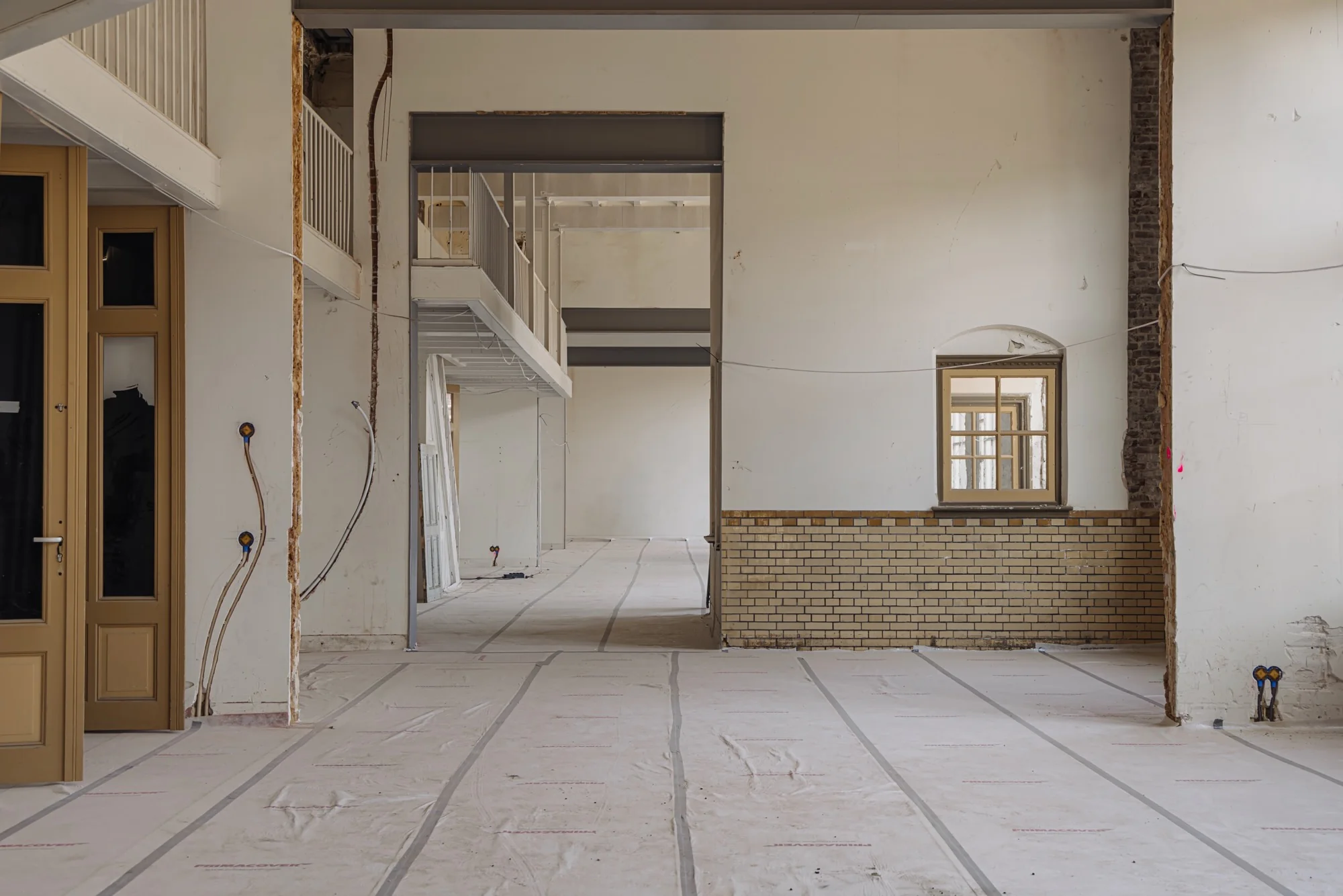 Interieur van een leegstaand pand aan de Leidseweg in verbouwing met witte muren, bakstenen lambrisering en zicht op een verdieping met balustrade.