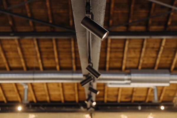 Zwarte spots op een plafondbalk in een industriële ruimte aan de Leidseweg met zicht op houten dakconstructie en ventilatiebuizen.