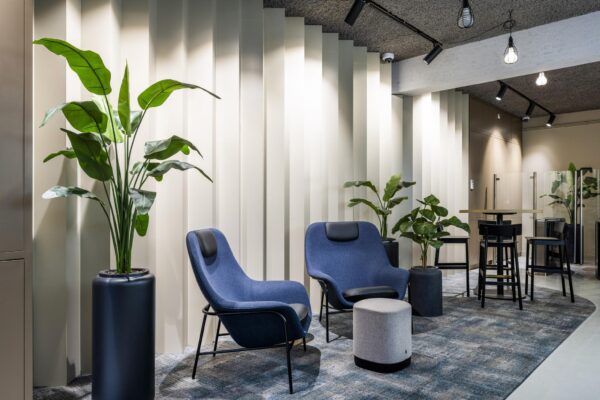Moderne wachtruimte met blauwe loungestoelen, hoge tafel met krukken, planten en een lichtgolvende wand.