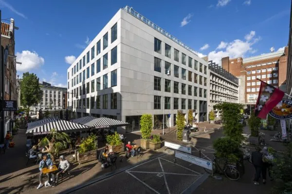 Terras van Vegan Junk Food Bar aan de Singel in Amsterdam met moderne kantoorgebouwen en voetgangers in de zon.
