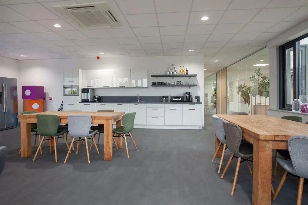 Moderne kantoorkeuken met houten eettafels, kleurrijke stoelen en een strak wit keukengedeelte aan de Cypresbaan.
