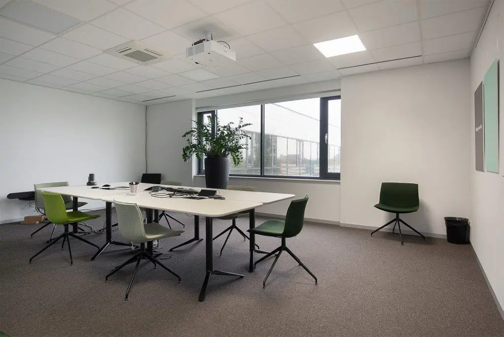 Vergaderruimte aan de Cypresbaan met witte tafel, groene stoelen, grote plant en plafondbeamer.