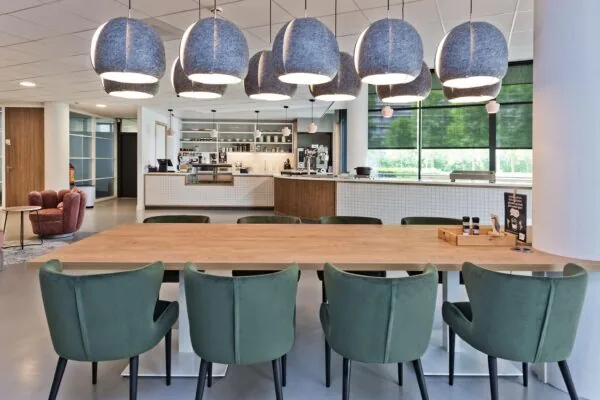 Moderne, open kantoorkantine met lange houten tafel, groene stoelen en hanglampen, aan de Prof. W.H. Keesomlaan.