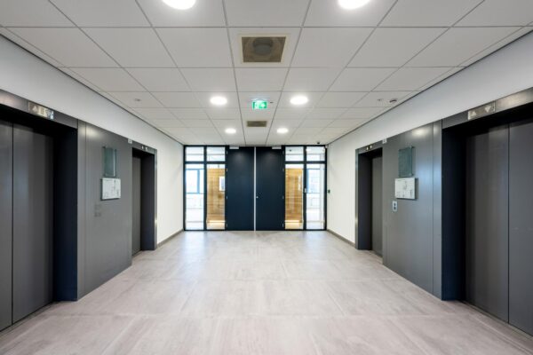 Moderne liftlobby met vier gesloten liften en een dubbele glazen deur aan het einde van de gang in een kantoorgebouw aan de Prof. W.H. Keesomlaan.