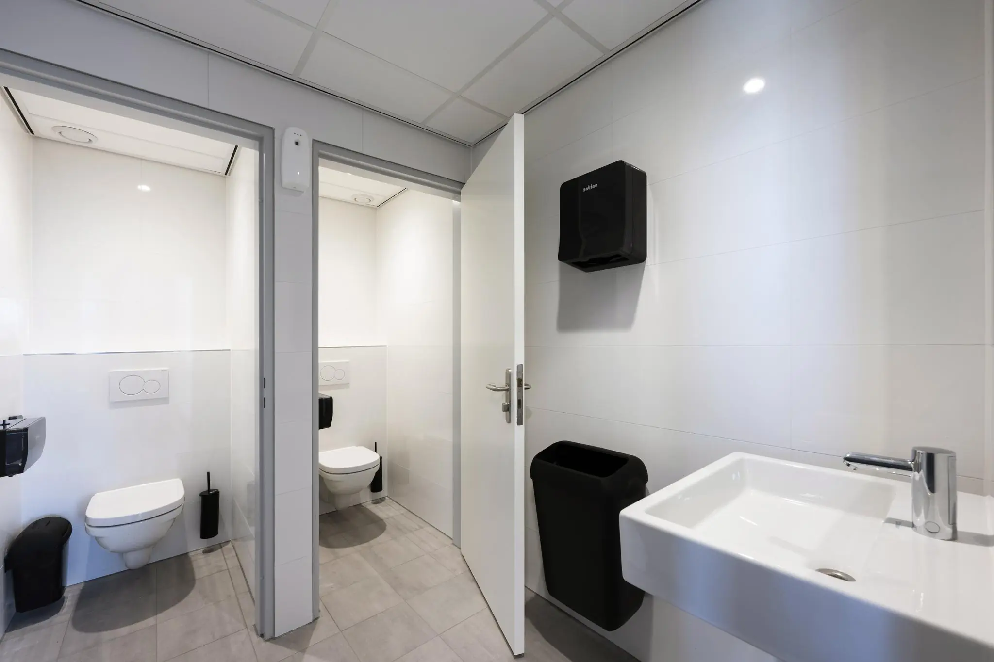 Moderne, wit betegelde toiletruimte met twee afgesloten toiletcabines, een wastafel en zwarte hygiënevoorzieningen aan de Prof. W.H. Keesomlaan.