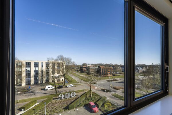 Uitzicht vanuit een raam op de Prof. W.H. Keesomlaan met kantoren, woonhuizen en een parkeerplaats onder een heldere blauwe lucht.