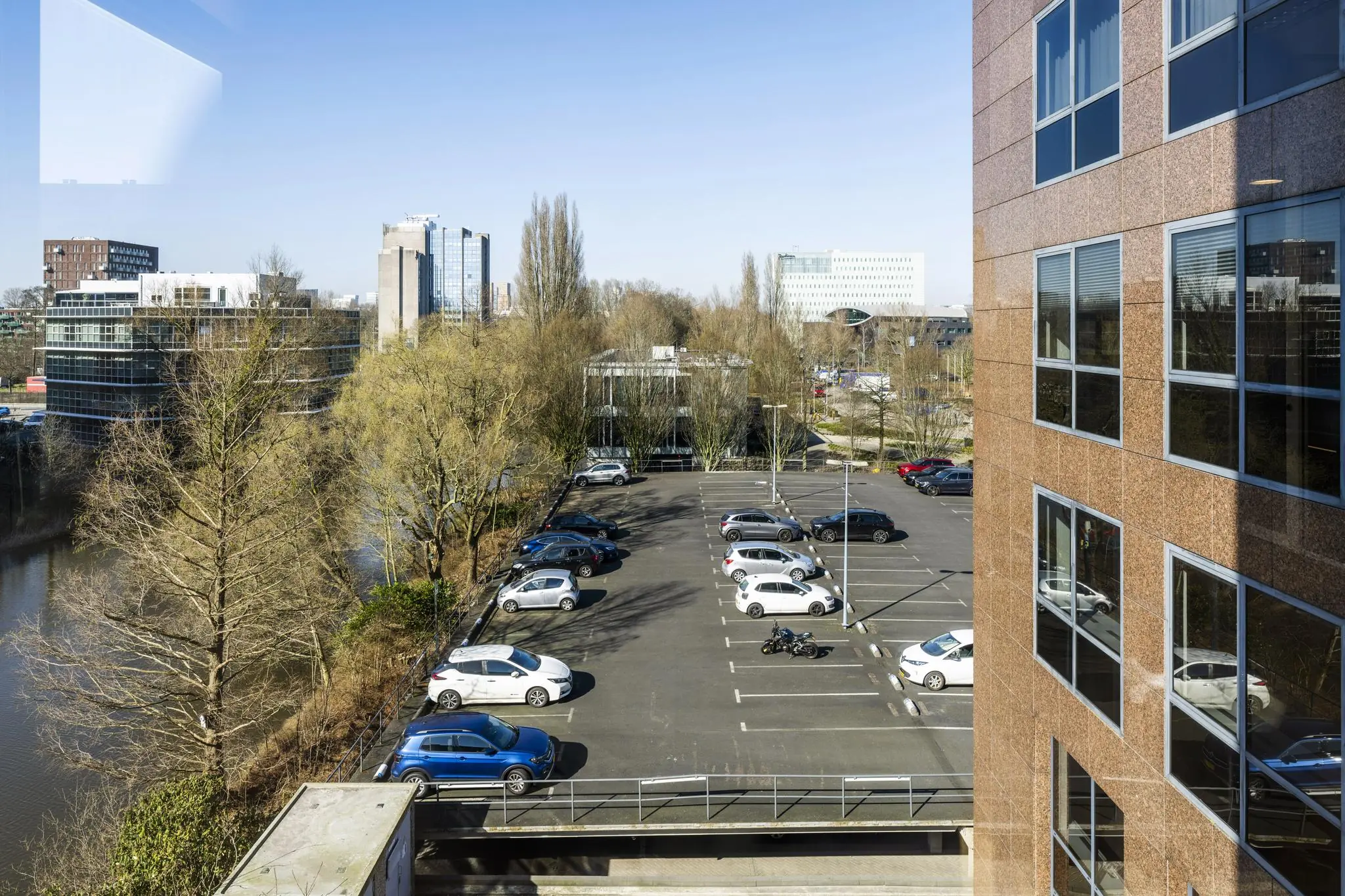 Zicht op een deels gevulde parkeerplaats aan de Prof. W.H. Keesomlaan met moderne kantoorgebouwen en bomen op de achtergrond.
