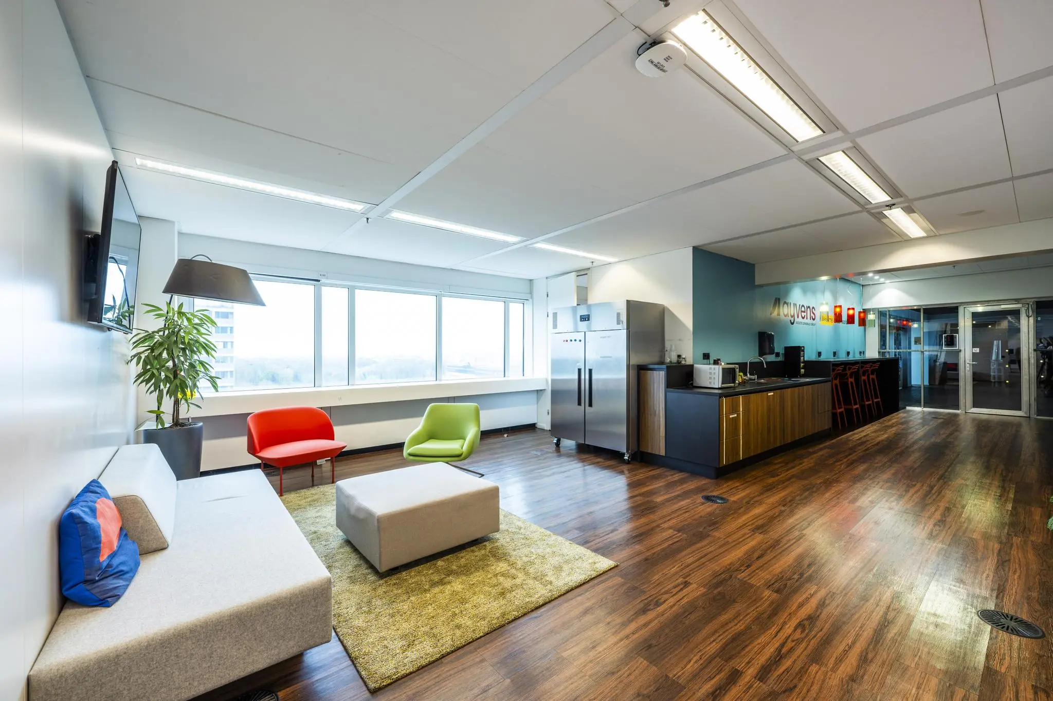 Moderne kantoorkeuken met loungehoek aan de Johan Huizingalaan, voorzien van kleurrijke stoelen, grote ramen en houten vloer.