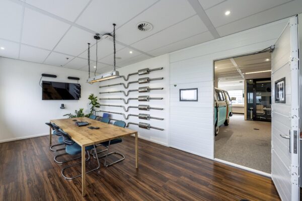 Vergaderruimte met houten tafel, wanddecoratie van uitlaten, en zicht op een klassieke auto in aangrenzende ruimte in een kantoor aan de Johan Huizingalaan.