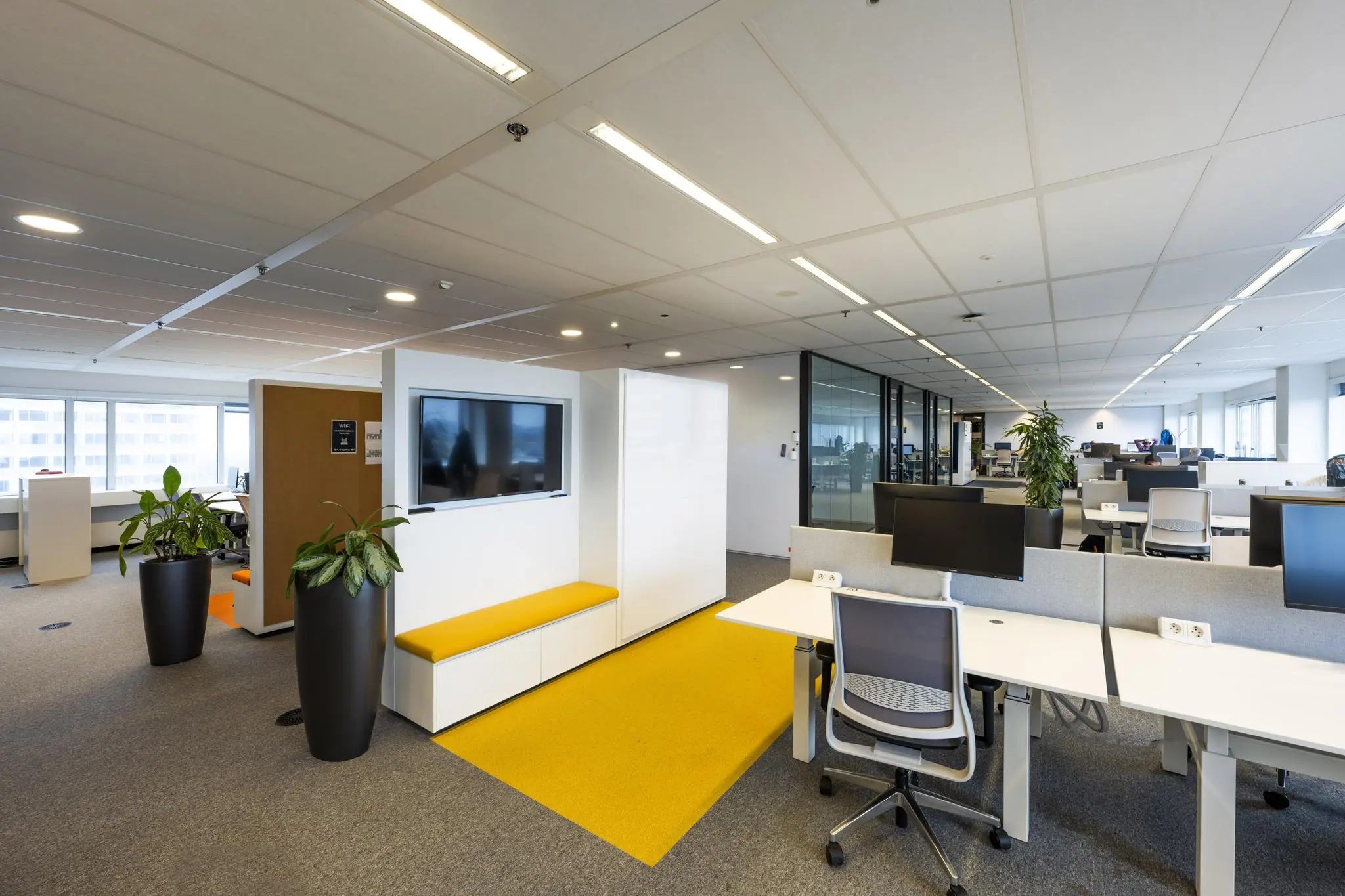 Modern kantoorinterieur aan de Johan Huizingalaan met flexwerkplekken, een gele loper en een zithoek met tv-scherm.