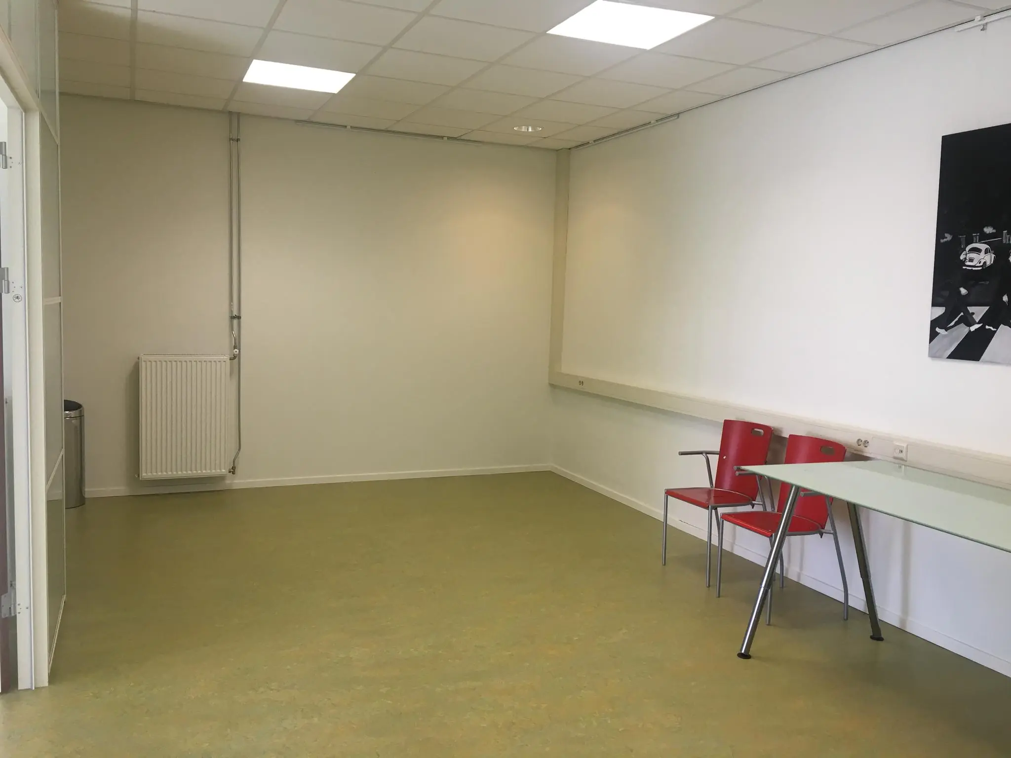 Lege wachtruimte met twee rode stoelen, een glazen tafel en een zwart-witfoto aan de muur in De Corridor.