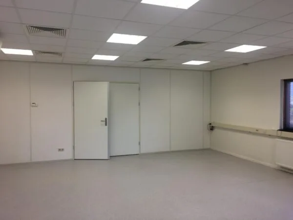 Lege, witte kantoorruimte met systeemplafond, twee gesloten deuren en een smal raam rechts.