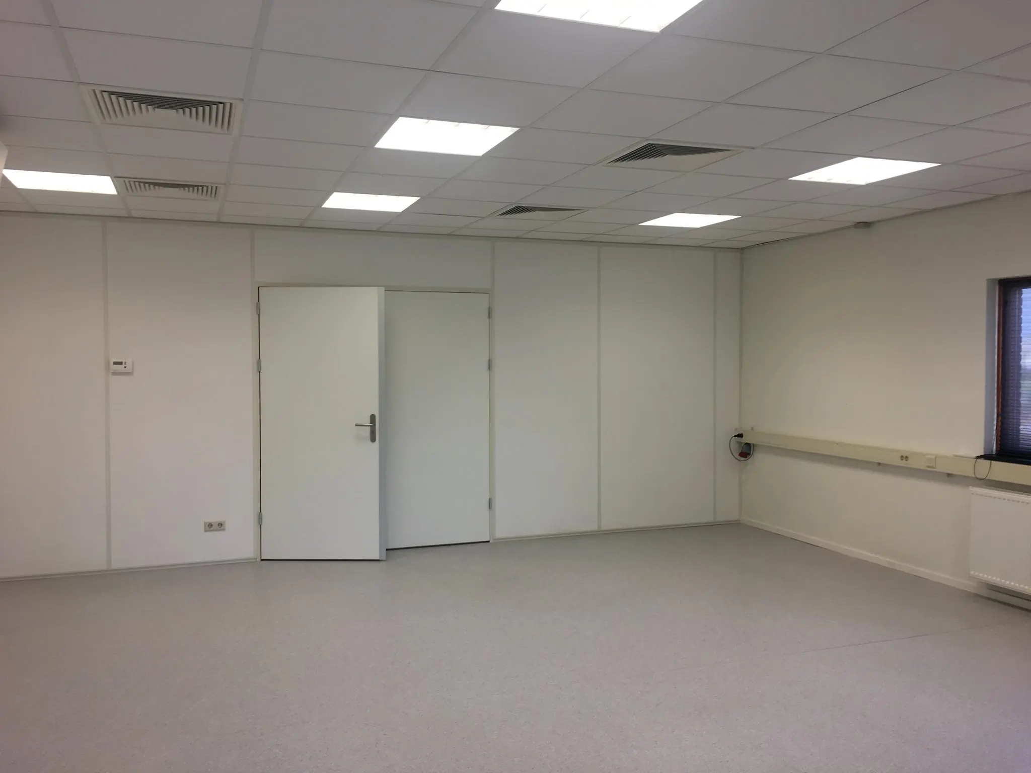 Lege, witte kantoorruimte met systeemplafond, twee gesloten deuren en een smal raam rechts.