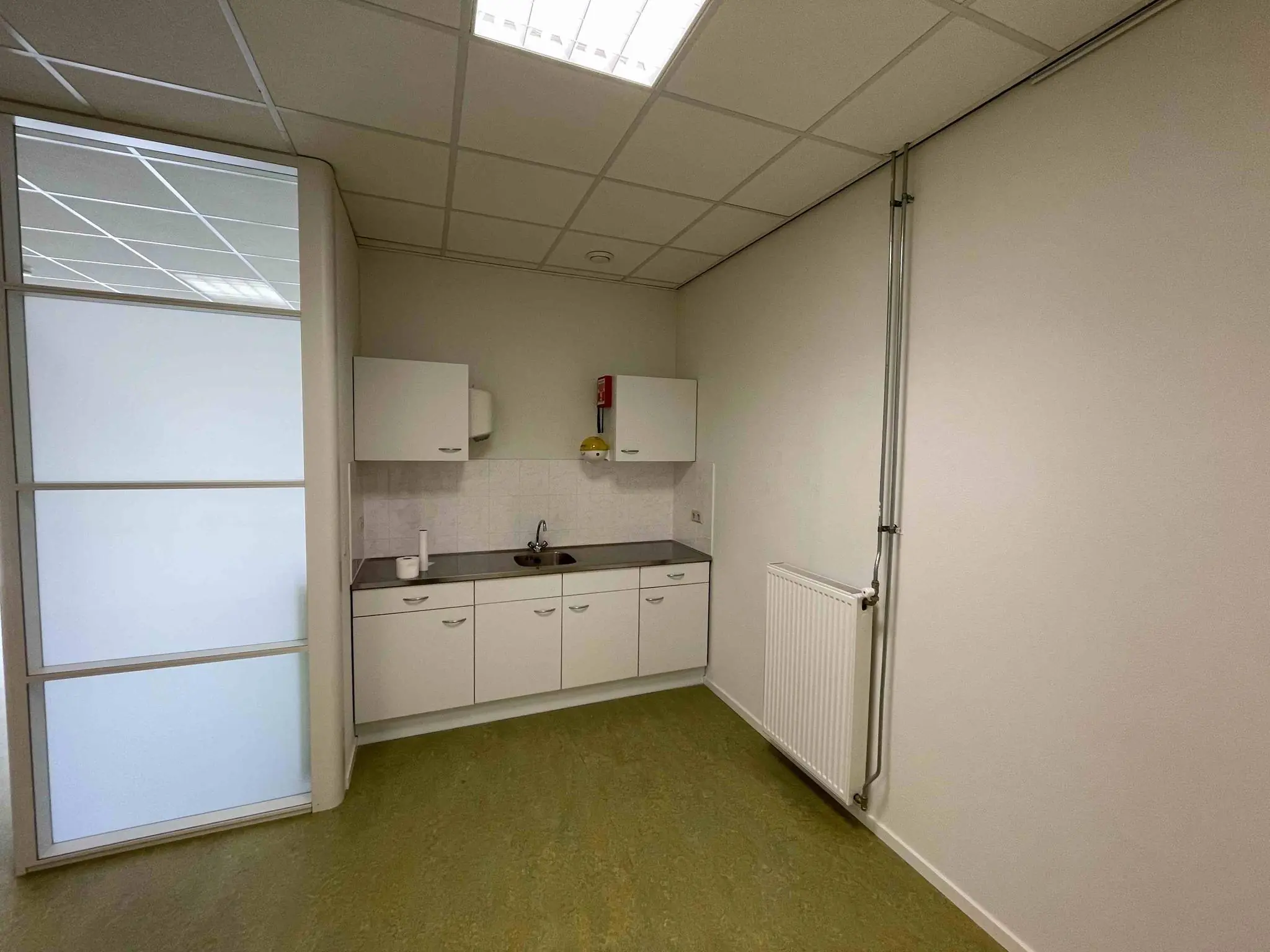 Kleine pantry met witte keukenkastjes, een spoelbak en een radiator in een kantoorruimte aan De Corridor.