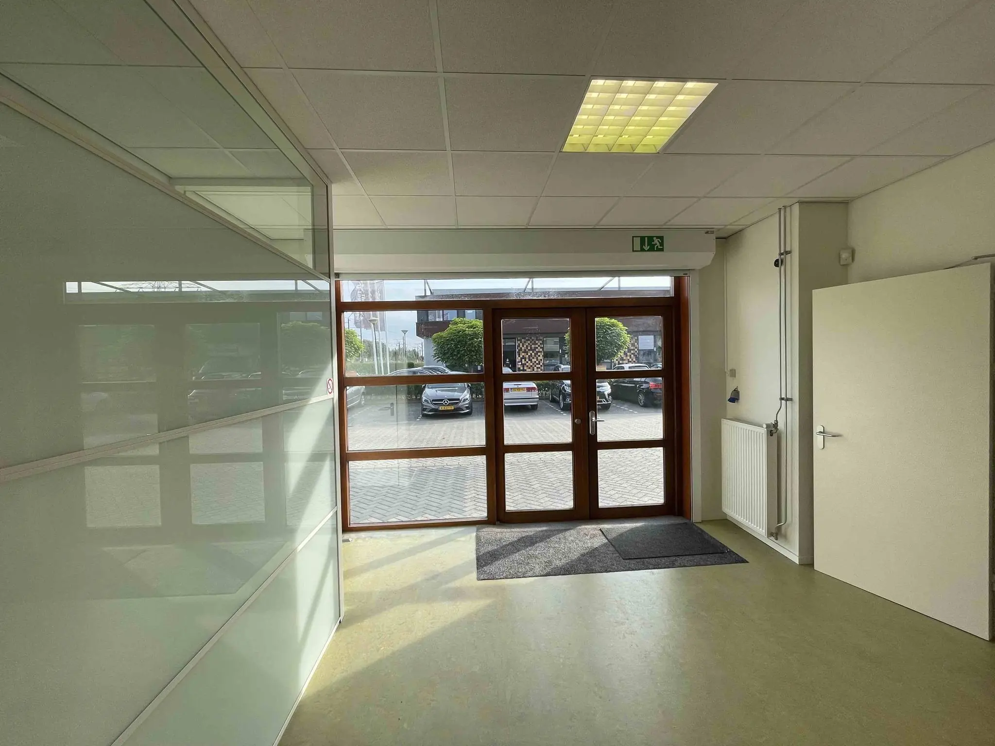 Inkomhal van De Corridor met glazen wand, open deur en zicht op geparkeerde auto's buiten.
