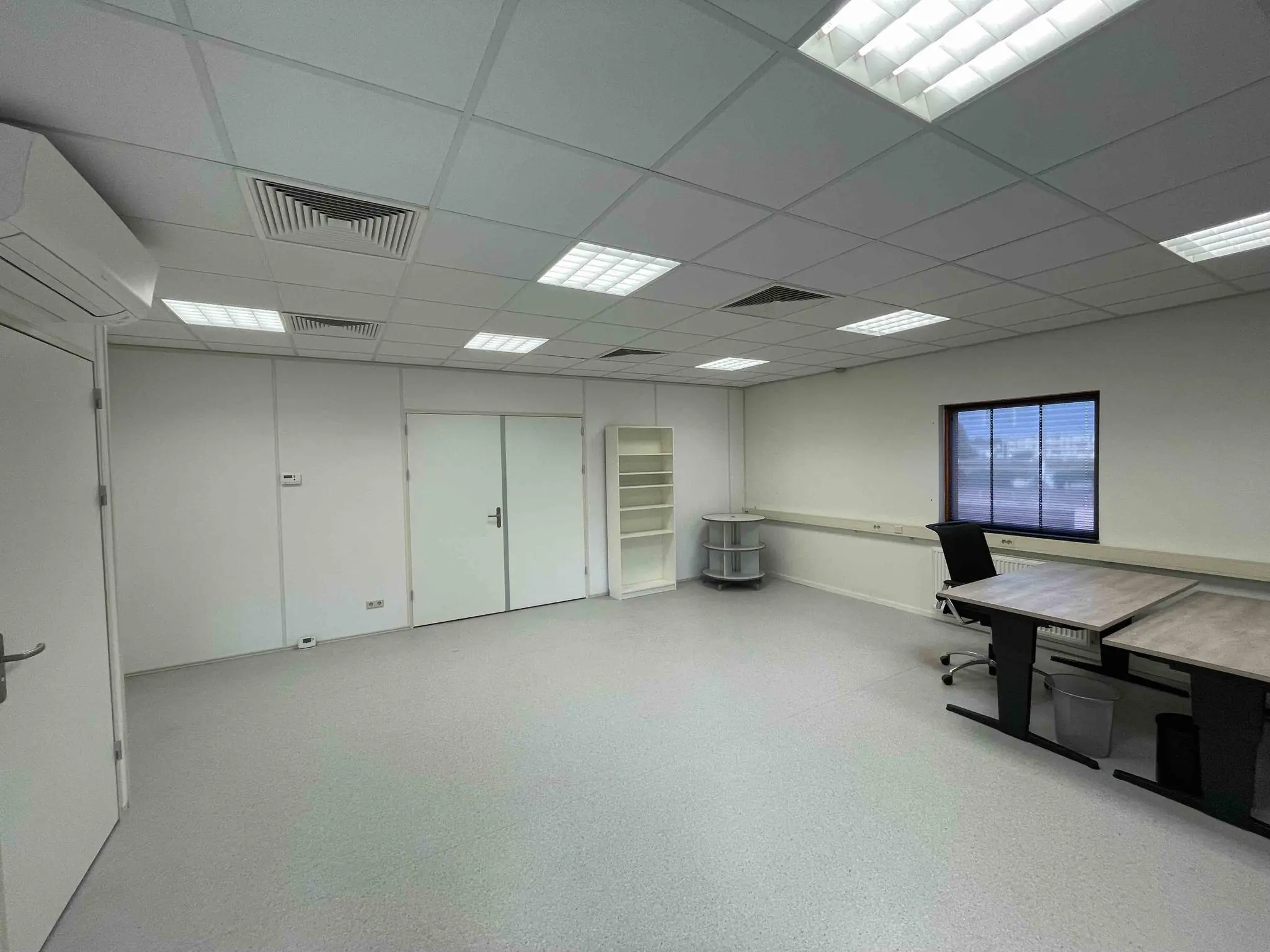 Kantoorruimte in De Corridor met bureaus, een stoel, opbergmeubels en een raam met zonwering.