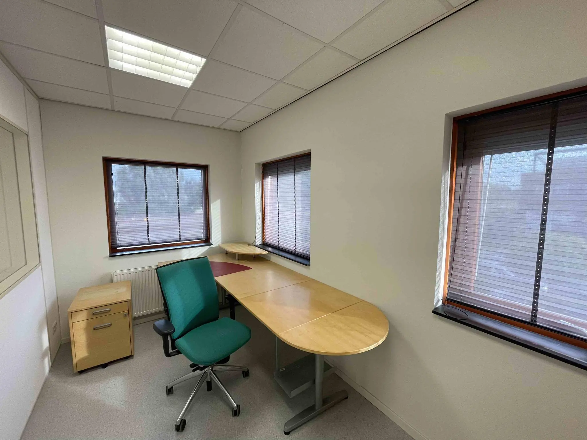 Klein kantoor met een houten bureau, groene bureaustoel en drie ramen met jaloezieën in bedrijfsverzamelgebouw De Corridor.