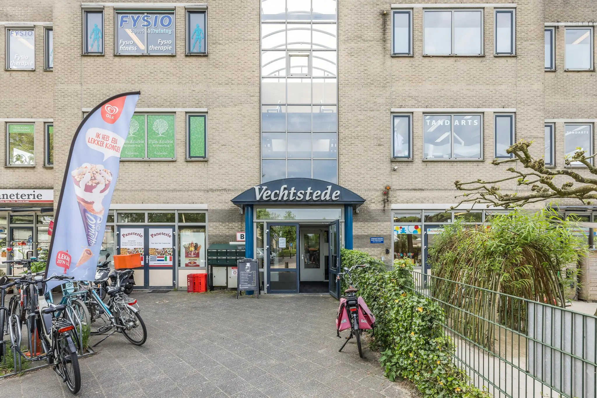 Ingang van gezondheidscentrum Vechtstede aan de Buitenweg met fietsen, winkels en een ijsreclamevlag op de voorgrond.