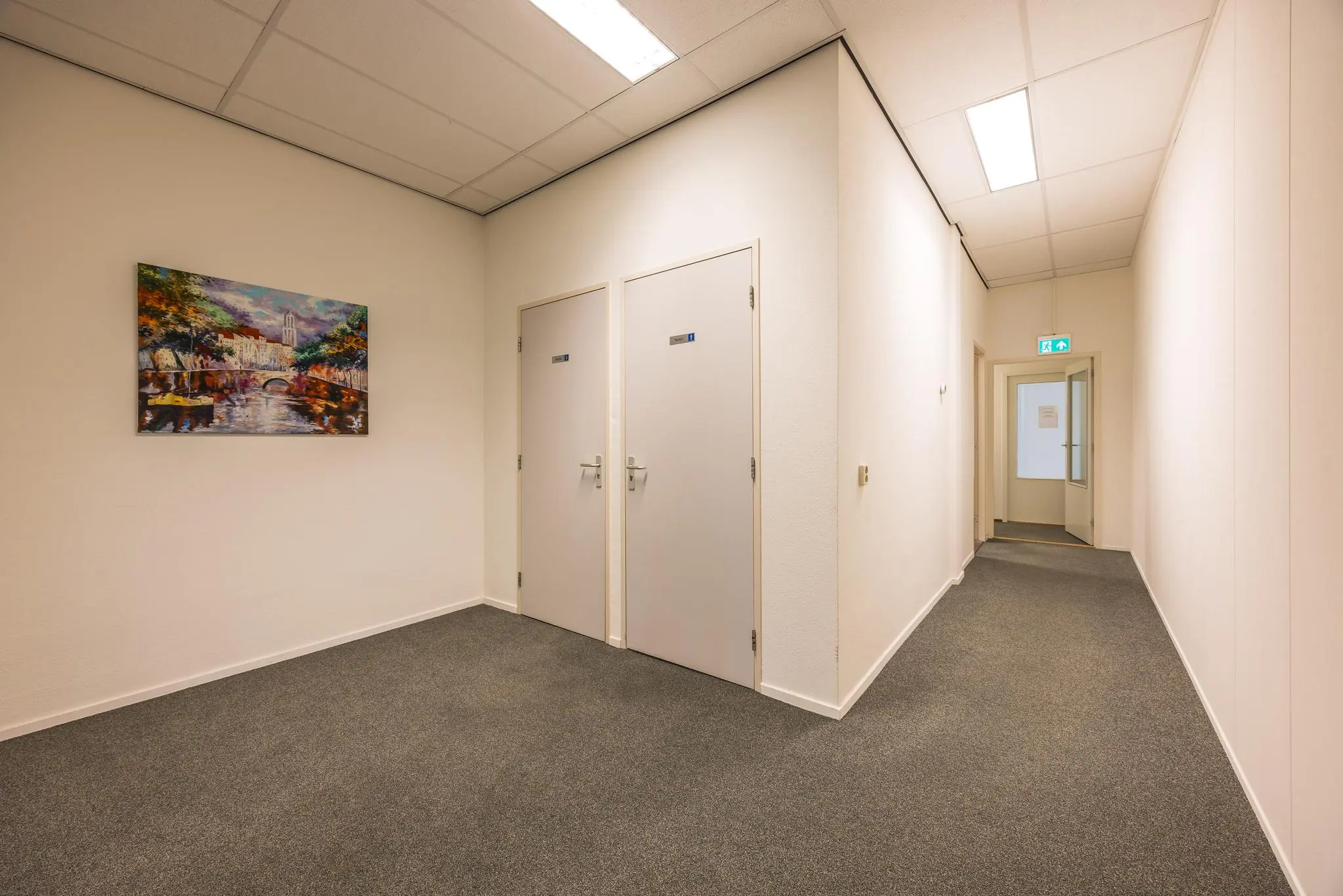 Verlichte gang in een kantoorruimte met twee toiletten, een schilderij aan de muur en een open deur aan het einde.