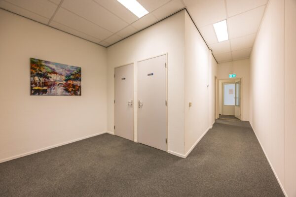 Verlichte gang met twee gesloten deuren, een schilderij aan de muur en een open deur naar een andere ruimte aan het einde van de gang.