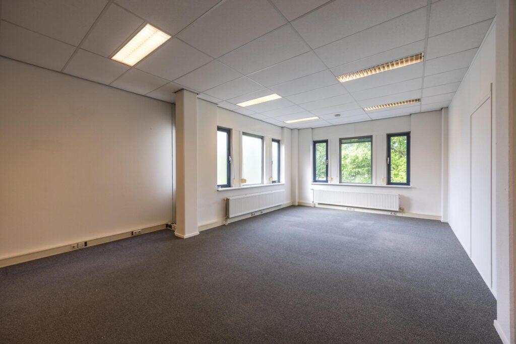 Lichte, lege kantoorruimte aan de Buitenweg met grijze vloerbedekking en meerdere ramen.